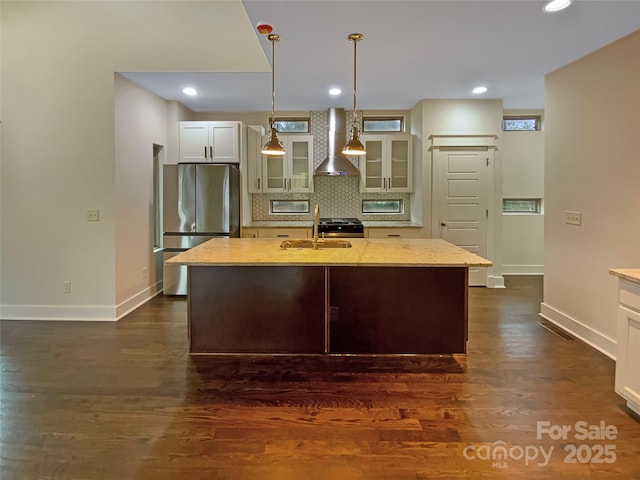 kitchen featuring an island with sink, glass insert cabinets, freestanding refrigerator, wall chimney range hood, and decorative backsplash