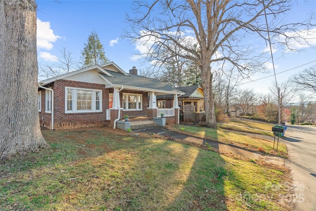 bungalow featuring a front yard, covered porch, a shingled roof, a chimney, and brick siding