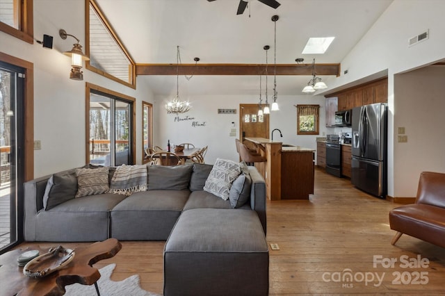 living area featuring ceiling fan with notable chandelier, light wood-style floors, visible vents, and high vaulted ceiling