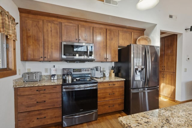 kitchen with light wood-type flooring, stainless steel appliances, brown cabinets, and light stone countertops