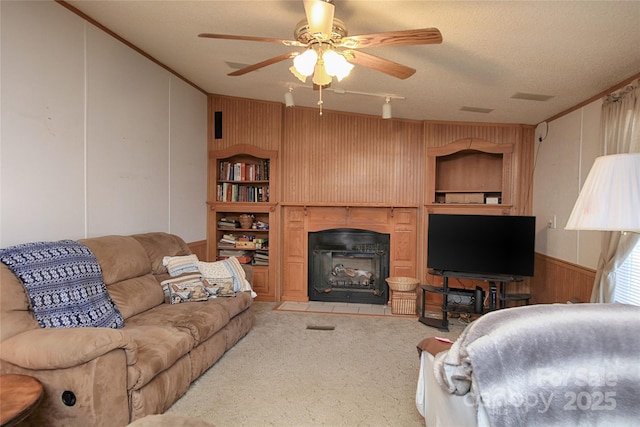 living room with crown molding, ceiling fan, a fireplace with flush hearth, wood walls, and light carpet