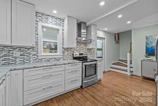 kitchen featuring light wood finished floors, stainless steel electric stove, decorative backsplash, white cabinetry, and wall chimney range hood