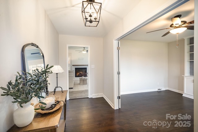 entryway featuring ceiling fan, baseboards, a warm lit fireplace, and dark wood finished floors