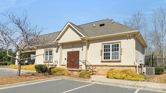 view of front of house featuring stucco siding, stone siding, fence, roof with shingles, and central AC unit