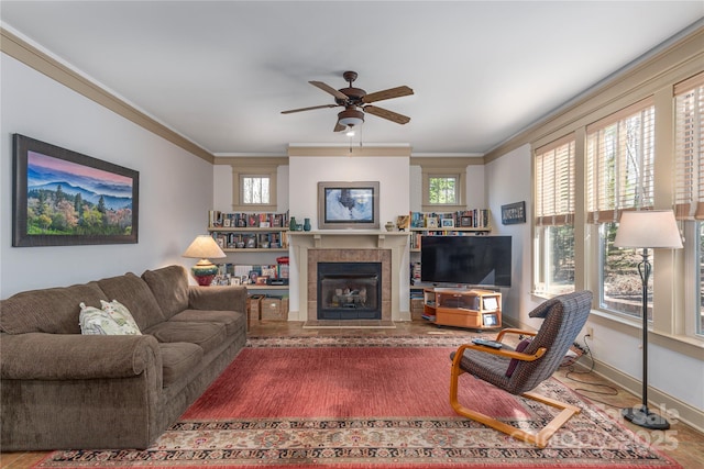 living area with a tiled fireplace, a ceiling fan, baseboards, and ornamental molding