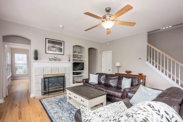 living room featuring built in features, arched walkways, a fireplace, light wood finished floors, and stairs