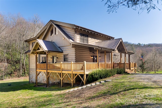 back of property with a ceiling fan, a wooden deck, a view of trees, a lawn, and metal roof