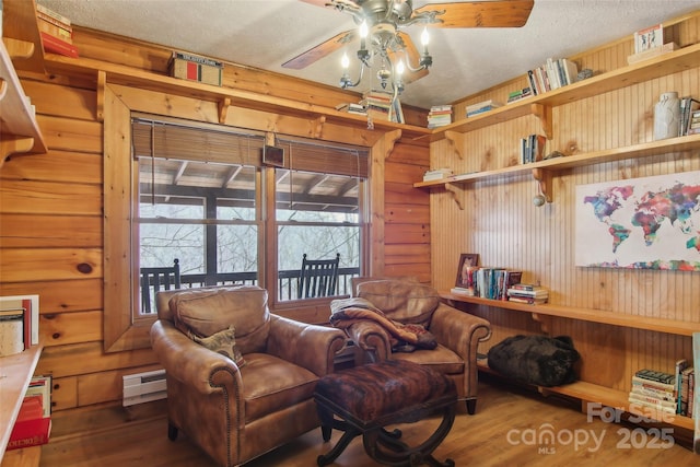 sitting room with wood walls, a baseboard heating unit, a ceiling fan, and wood finished floors