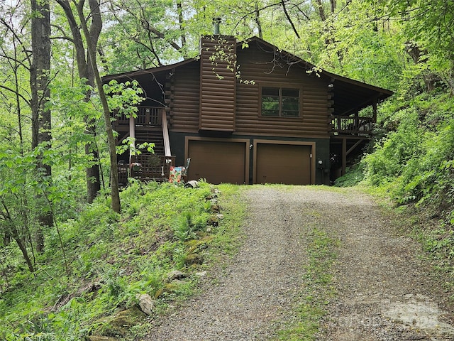 view of front of house featuring log siding, an attached garage, dirt driveway, a chimney, and a wooded view
