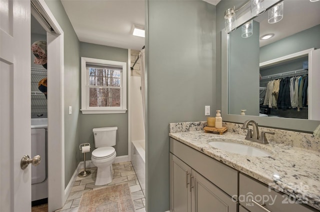 bathroom featuring a walk in closet, baseboards, toilet, stone tile floors, and vanity