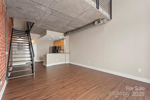 unfurnished living room with brick wall, stairs, dark wood-type flooring, and baseboards