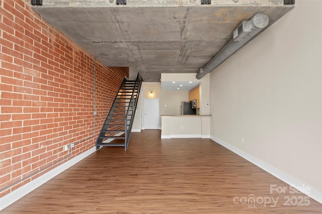 unfurnished living room featuring stairway, wood finished floors, baseboards, and brick wall