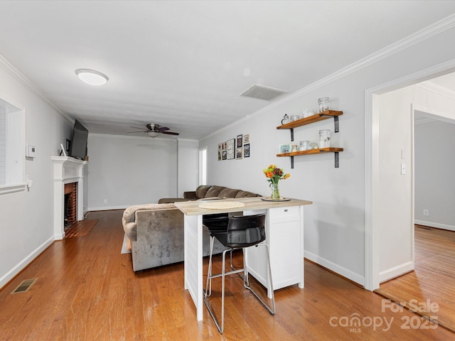 living area featuring a ceiling fan, visible vents, a fireplace with flush hearth, ornamental molding, and light wood-type flooring