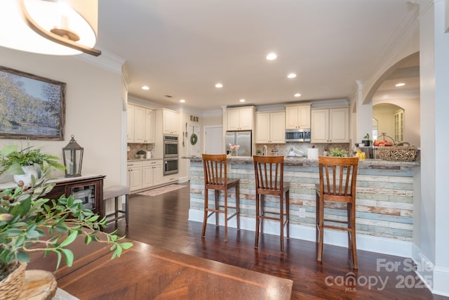 kitchen with dark wood-type flooring, a breakfast bar, ornamental molding, arched walkways, and appliances with stainless steel finishes