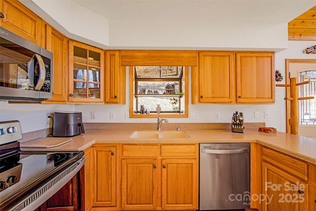 kitchen with a sink, stainless steel appliances, brown cabinetry, light countertops, and glass insert cabinets