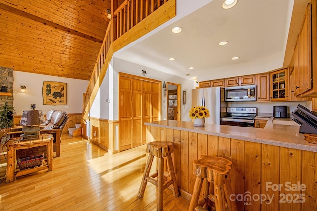 kitchen featuring a wainscoted wall, light wood-style floors, appliances with stainless steel finishes, a peninsula, and brown cabinetry