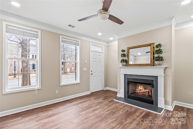 unfurnished living room with visible vents, ornamental molding, a warm lit fireplace, wood finished floors, and baseboards