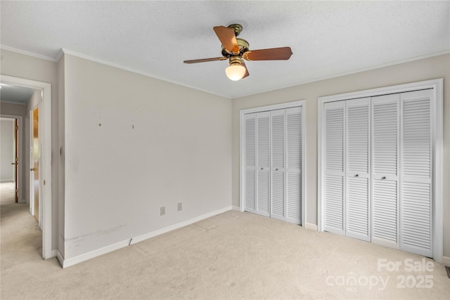 unfurnished bedroom with baseboards, a textured ceiling, crown molding, light colored carpet, and two closets