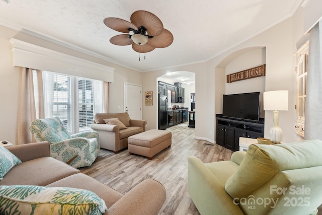 living area featuring a ceiling fan, baseboards, light wood-style flooring, arched walkways, and ornamental molding