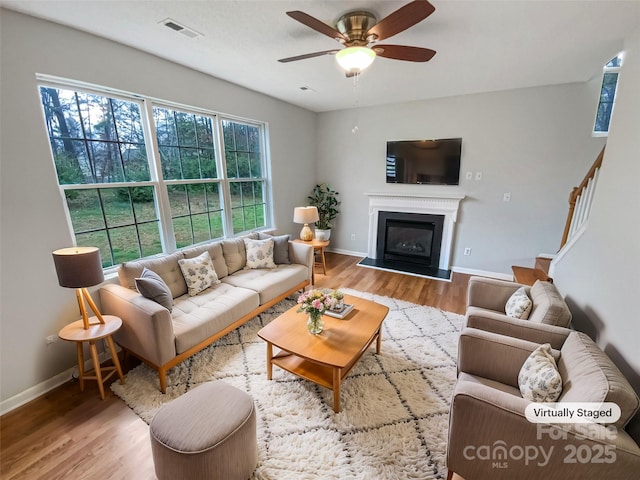 living area featuring visible vents, baseboards, wood finished floors, a glass covered fireplace, and a ceiling fan