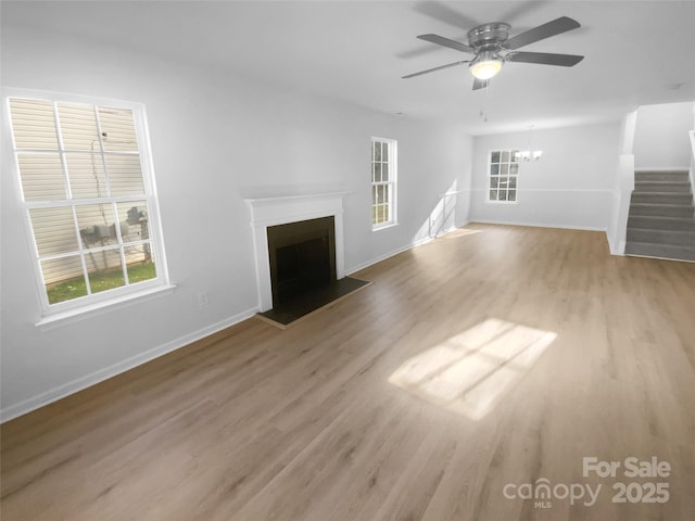 unfurnished living room featuring light wood-type flooring, ceiling fan with notable chandelier, stairway, a fireplace, and baseboards