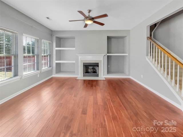 unfurnished living room with built in shelves, baseboards, a fireplace with flush hearth, stairs, and wood finished floors