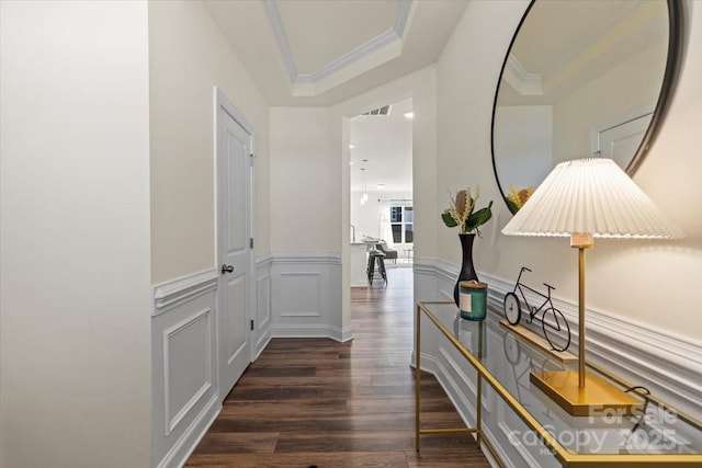 hallway featuring ornamental molding, wainscoting, a decorative wall, a raised ceiling, and dark wood-style flooring