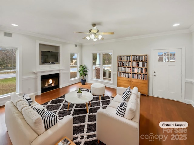 living room featuring recessed lighting, a warm lit fireplace, crown molding, and wood finished floors