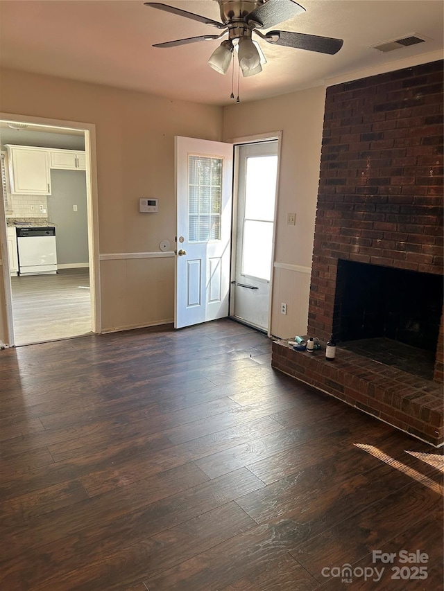 unfurnished living room featuring dark wood-style floors, a wainscoted wall, visible vents, a ceiling fan, and a brick fireplace