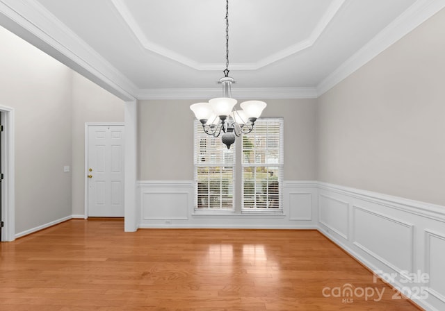 unfurnished dining area with a wainscoted wall, a notable chandelier, ornamental molding, a tray ceiling, and light wood-style floors