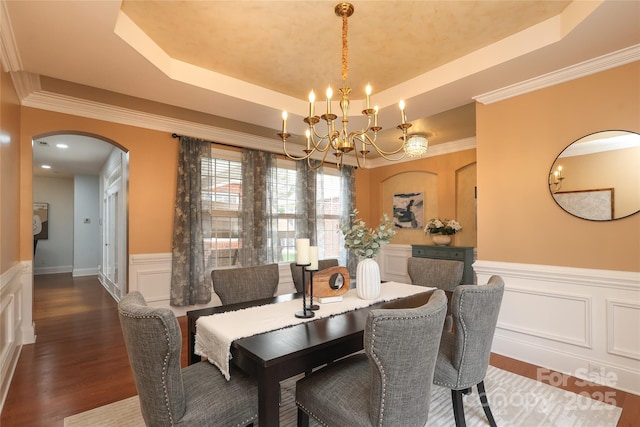 dining space with a wainscoted wall, wood finished floors, arched walkways, an inviting chandelier, and a raised ceiling