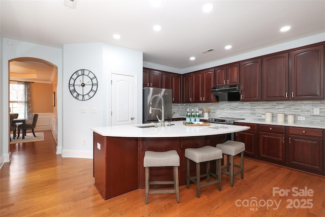 kitchen featuring an island with sink, arched walkways, freestanding refrigerator, light countertops, and light wood-type flooring