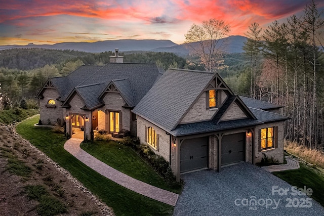 french provincial home featuring a wooded view, gravel driveway, a front yard, a chimney, and a mountain view