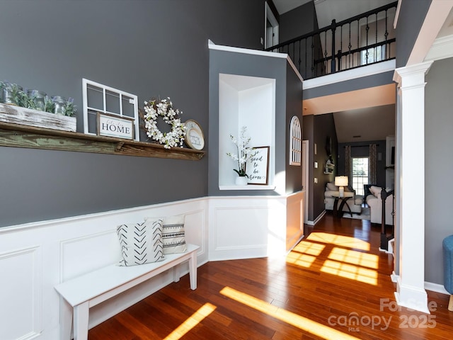 interior space featuring a wainscoted wall, wood finished floors, a decorative wall, a towering ceiling, and ornate columns