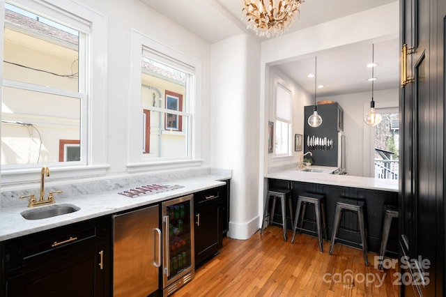 kitchen with beverage cooler, a sink, decorative light fixtures, dark cabinetry, and light wood-style floors