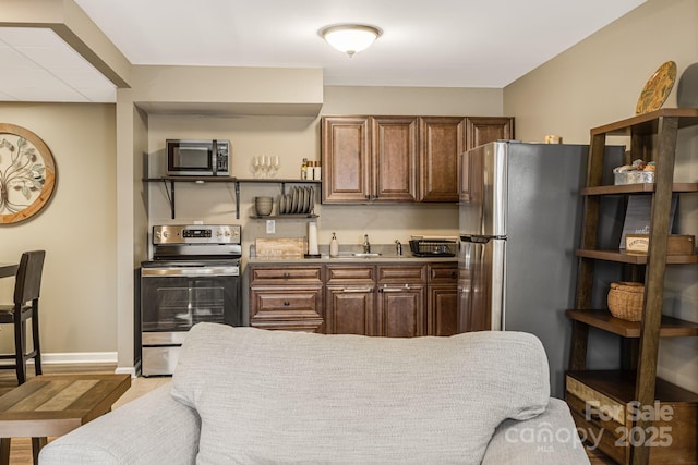 kitchen featuring open shelves, a sink, stainless steel appliances, brown cabinetry, and baseboards