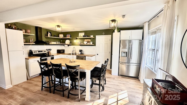 kitchen with open shelves, appliances with stainless steel finishes, wall chimney exhaust hood, and white cabinets