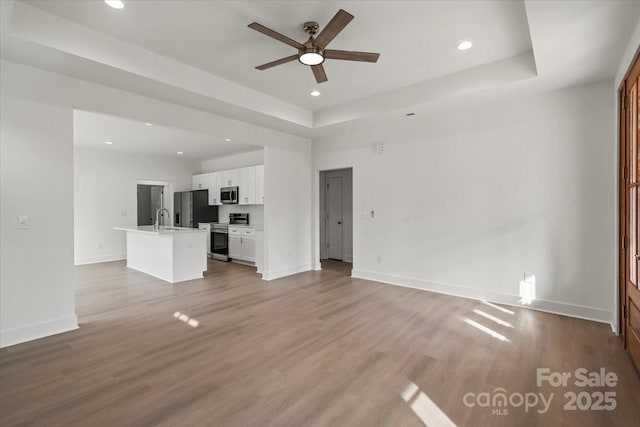 unfurnished living room featuring a tray ceiling, a ceiling fan, light wood-style floors, and a sink