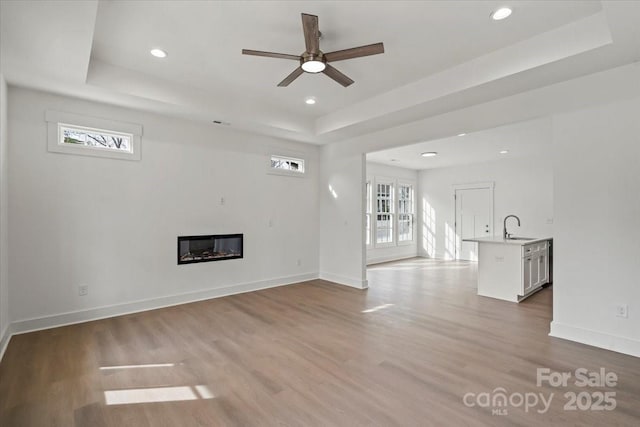 unfurnished living room featuring wood finished floors, baseboards, a tray ceiling, ceiling fan, and a sink