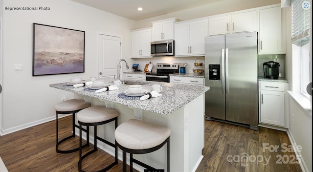 kitchen with dark wood-type flooring, a center island with sink, appliances with stainless steel finishes, a kitchen breakfast bar, and white cabinetry