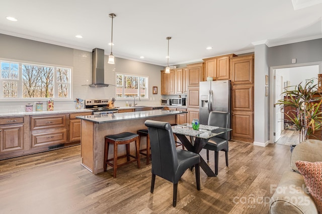 kitchen with wood finished floors, a kitchen island, appliances with stainless steel finishes, crown molding, and wall chimney range hood