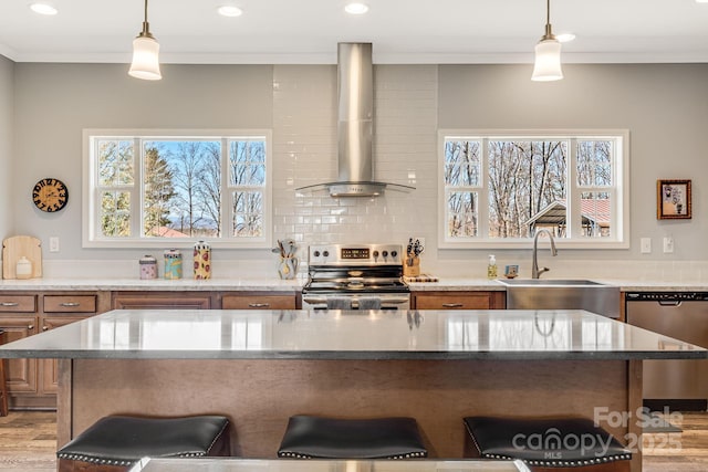 kitchen with brown cabinets, a breakfast bar, a sink, appliances with stainless steel finishes, and wall chimney exhaust hood