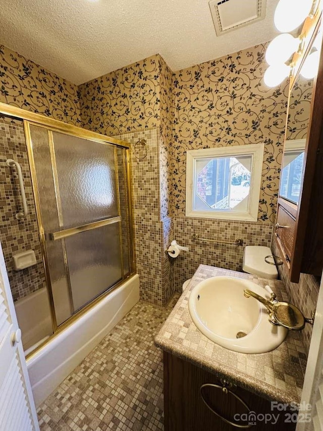 full bath with visible vents, a textured ceiling, combined bath / shower with glass door, wallpapered walls, and vanity