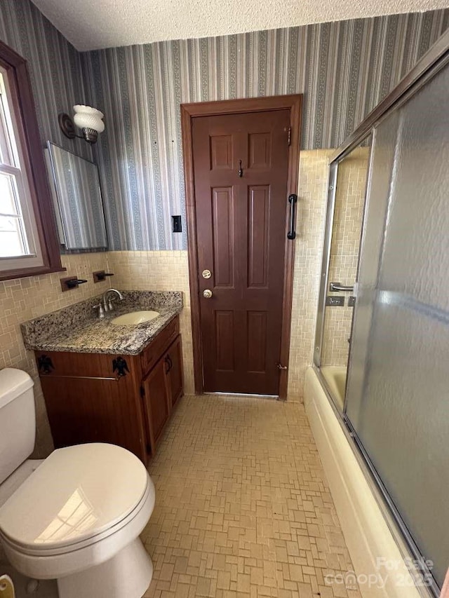 bathroom with toilet, a textured ceiling, combined bath / shower with glass door, wallpapered walls, and vanity