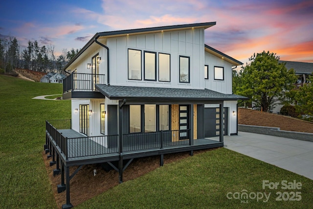 back of house at dusk with board and batten siding, roof with shingles, a lawn, a balcony, and driveway