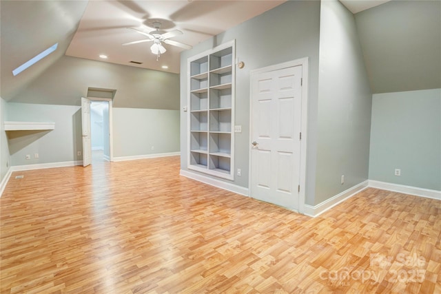 bonus room featuring built in shelves, a ceiling fan, vaulted ceiling with skylight, light wood-style floors, and baseboards