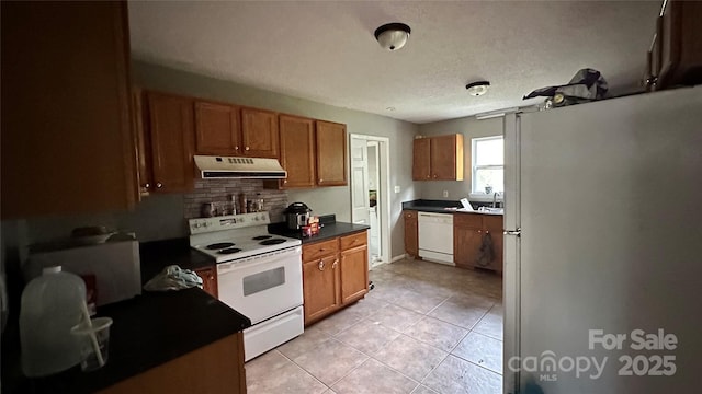 kitchen featuring brown cabinets, under cabinet range hood, dark countertops, white appliances, and light tile patterned floors