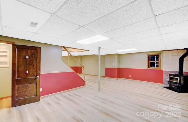 finished below grade area featuring wood finished floors, baseboards, visible vents, a wood stove, and a paneled ceiling