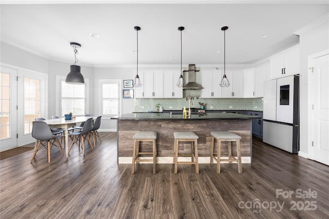 kitchen with tasteful backsplash, dark countertops, freestanding refrigerator, crown molding, and wall chimney range hood