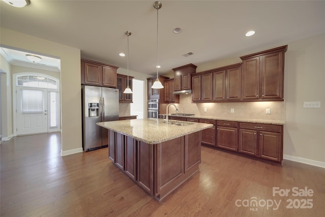 kitchen with dark wood-type flooring, light stone counters, decorative backsplash, appliances with stainless steel finishes, and a kitchen island with sink